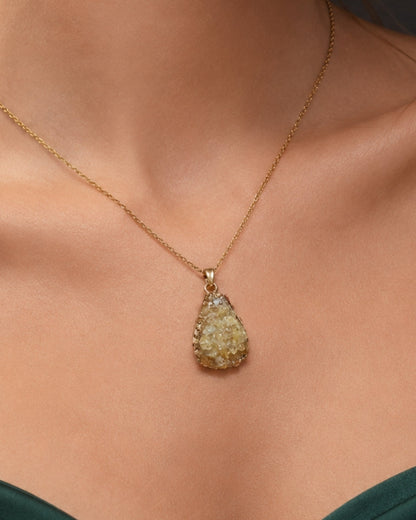 A gold teardrop pendant necklace with a textured, rough-hewn stone pendant, shown worn on a model's neck above a dark green satin top.