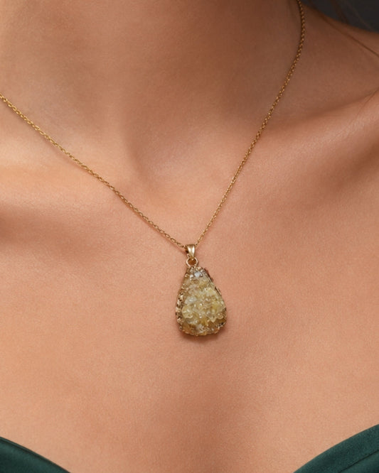 A gold teardrop pendant necklace with a textured, rough-hewn stone pendant, shown worn on a model's neck above a dark green satin top.