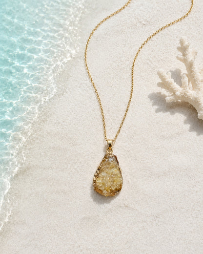 A gold teardrop pendant necklace with a textured, glittery yellow stone lies on white sand next to the edge of turquoise water and a white coral fragment.