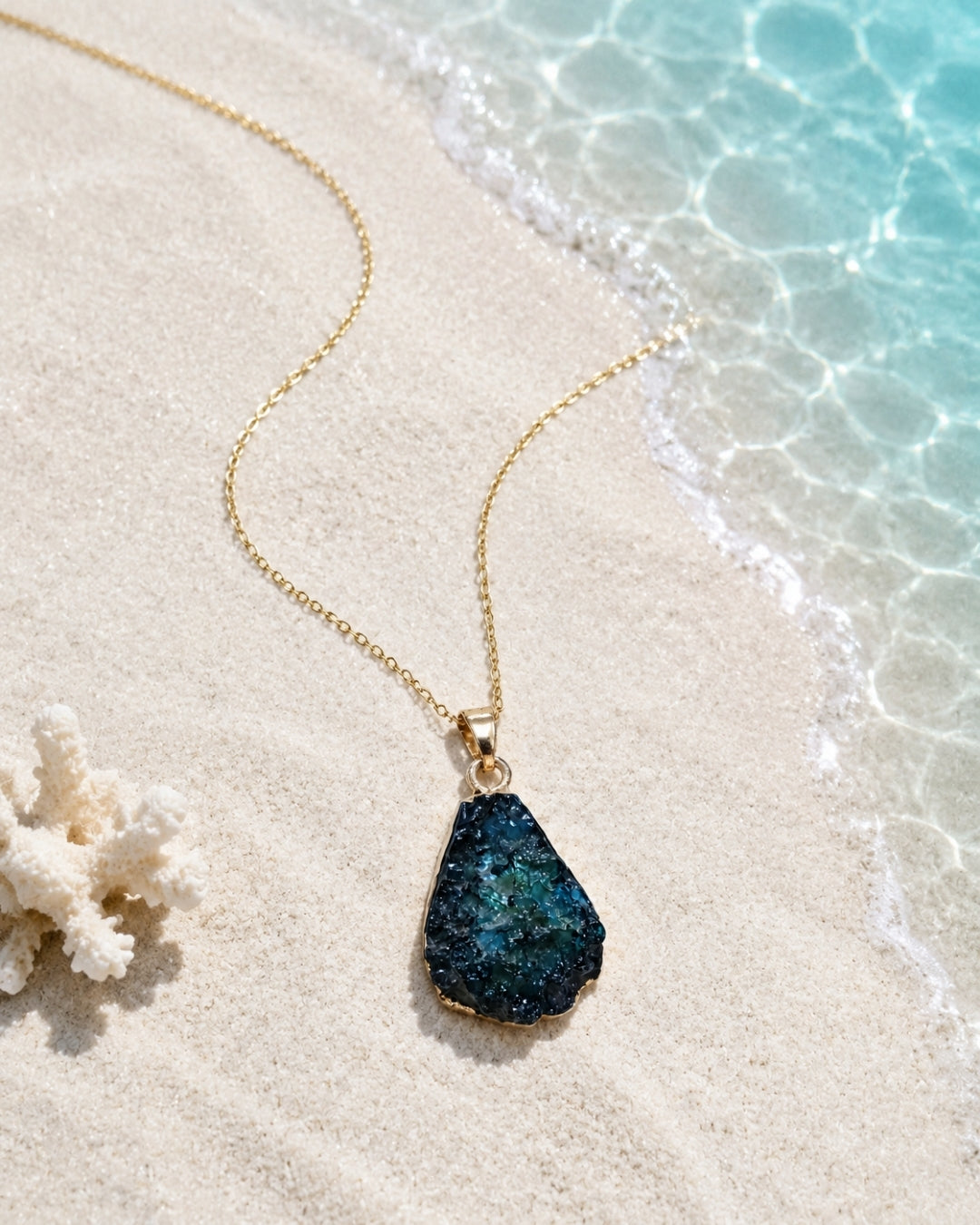 A gold chain necklace with a teardrop-shaped dark blue and green glittery druzy stone pendant, displayed on a sandy beach next to clear ocean water and a piece of coral.