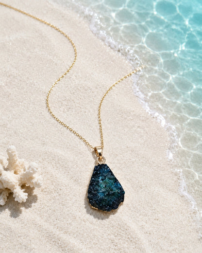 A gold chain necklace with a teardrop-shaped dark blue and green glittery druzy stone pendant, displayed on a sandy beach next to clear ocean water and a piece of coral.