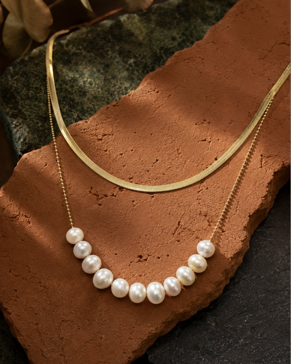 A layered necklace set displayed on a terracotta tile and dark stone background. The set includes a gold chain with a curved row of white freshwater pearls and a second gold chain with a smooth, curved gold pendant.