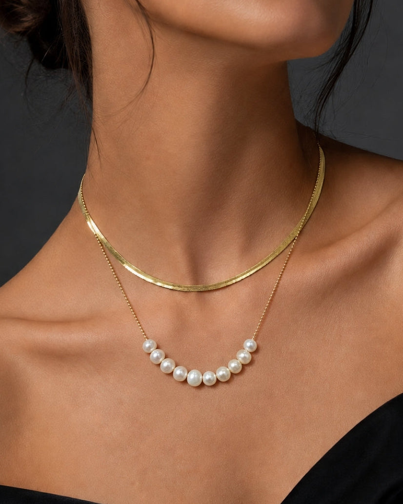 A close-up of a model wearing a double-layered necklace. The top layer is a simple polished gold chain, and the bottom layer is a delicate gold chain featuring a curved row of white freshwater pearls.
