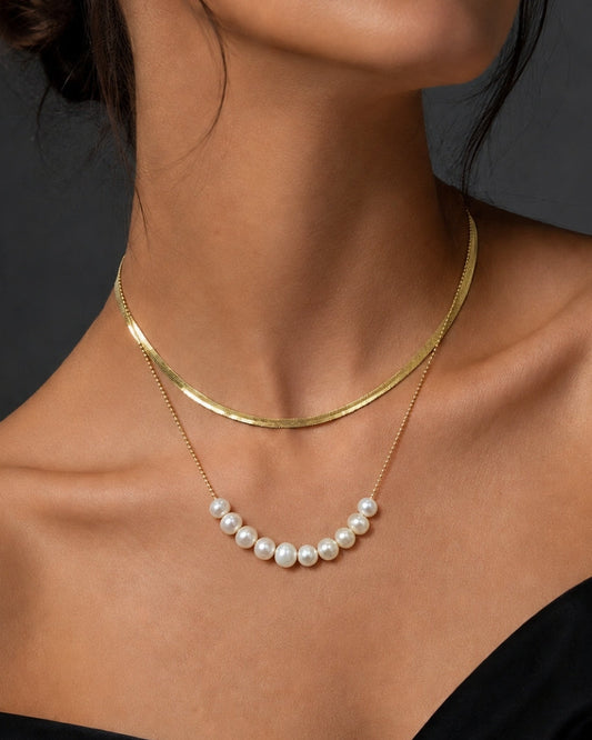 A close-up of a model wearing a double-layered necklace. The top layer is a simple polished gold chain, and the bottom layer is a delicate gold chain featuring a curved row of white freshwater pearls.