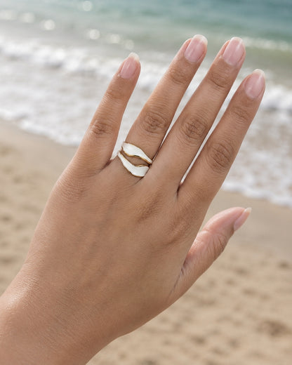 A close-up of a hand wearing a double-shell ring on the ring finger, set against a blurred beach background with ocean waves and sand.