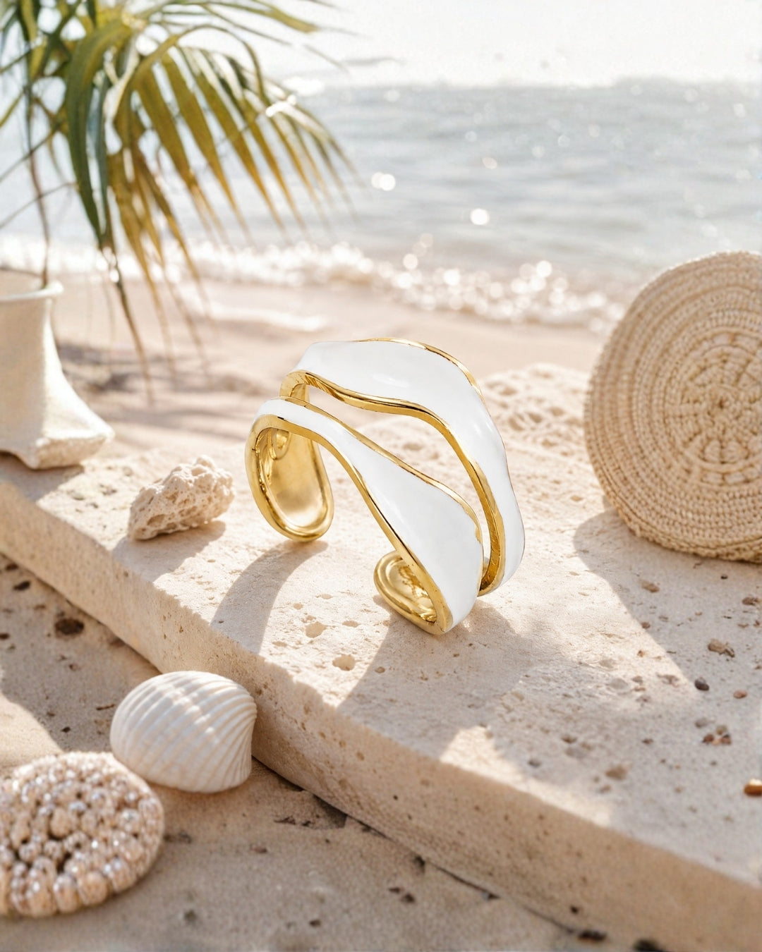 A wide, sculptural cuff bracelet with a glossy white surface and polished gold edges, displayed on a sandy beach next to seashells, a woven hat, and a palm leaf.